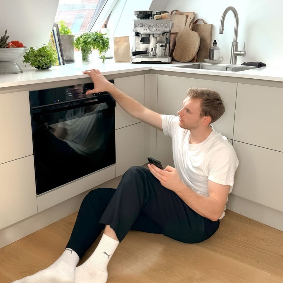 Person sitting on wooden floor in white kitchen with black oven | HTH