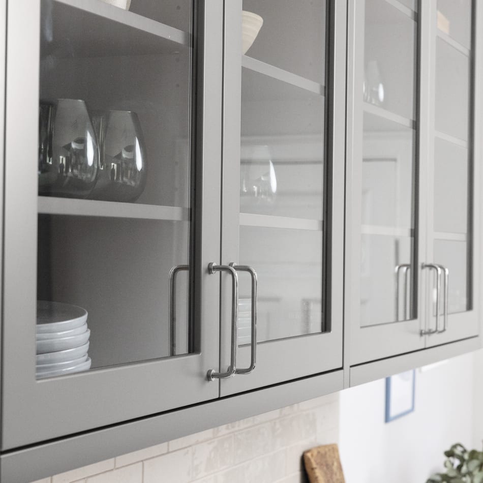 Gray kitchen cabinets with glass doors and metal handles above a light-colored countertop. Model Art | HTH