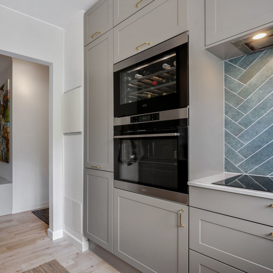 Gray kitchen cabinets with built-in oven and blue herringbone tile backsplash. Model Art | HTH