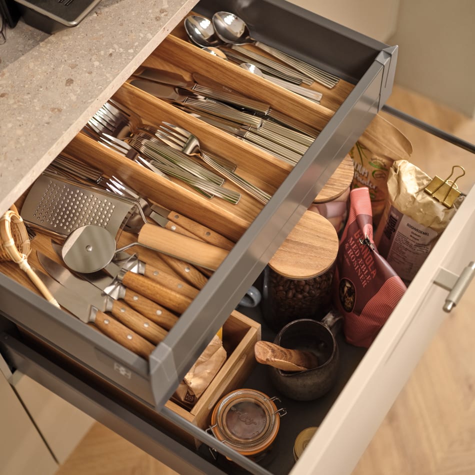 Open kitchen drawer with wooden dividers and utensils, light-colored countertop above. Reflection | HTH