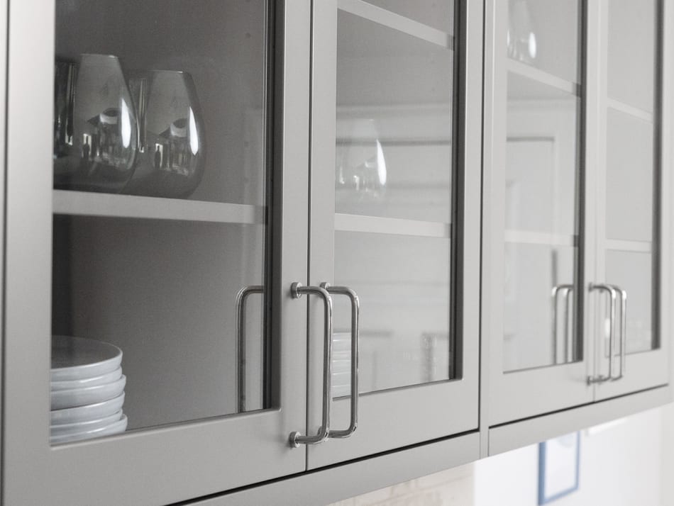 Gray kitchen cabinets with glass doors and metal handles above a light-colored countertop. Model Art | HTH